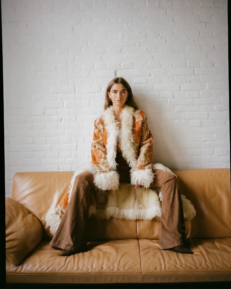 Editorial portrait — model in a shearling coat on the leather sofa, white brick behind